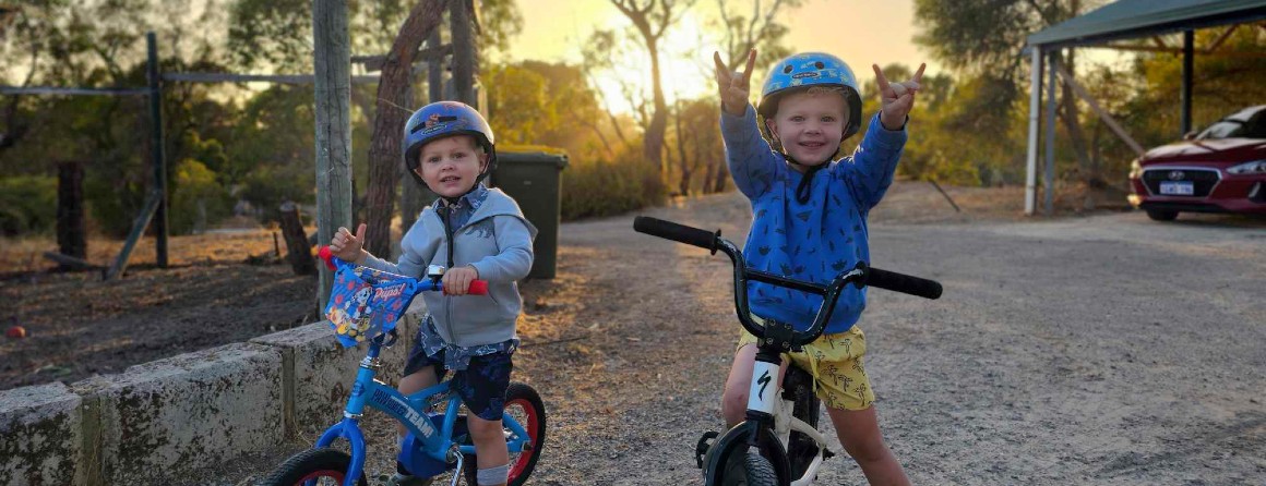 Two kinder-age brothers with blond hair ride on their bikes during sunset. Both are wearing helmets and one is gesturing to the camera.